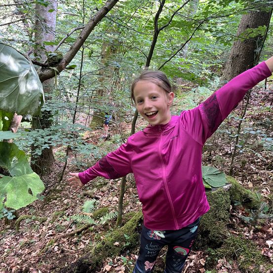 Ein junges Mädchen in einer pinkfarbenen Jacke lächelt und posiert für ein Foto im Wald. Sie steht auf einem moosbedeckten Felsen mit ausgestreckten Armen.