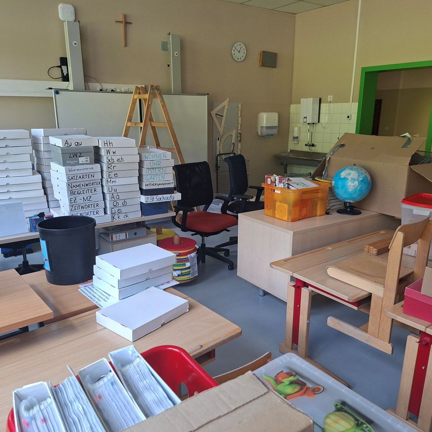 Ein Klassenzimmer mit mehreren Schreibtischen, Stühlen und Lagerboxen. Ein Schreibtisch hat eine Weltkugel und eine offene Box, während ein anderer einen Eimer hat. Stapel von beschrifteten Kisten stehen links, und eine Leiter lehnt an der Wand. Eine Uhr und ein Kreuz sind an der Wand montiert.