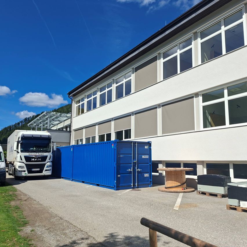 Ein großer blauer Seecontainer steht vor einem weißen Gebäude mit vielen Fenstern an einem sonnigen Tag.