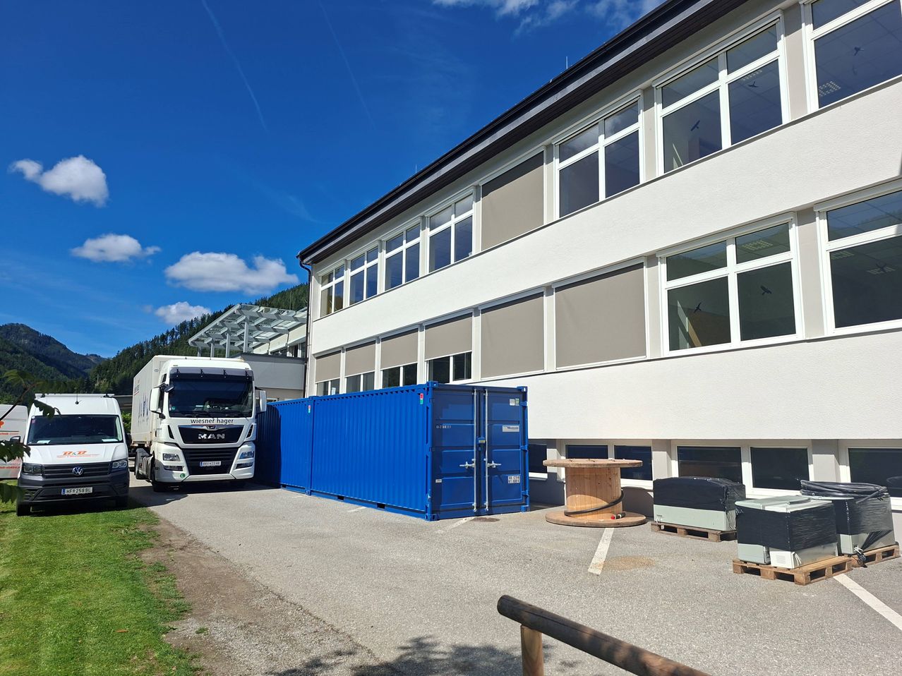Ein großer blauer Seecontainer steht vor einem weißen Gebäude mit vielen Fenstern an einem sonnigen Tag.