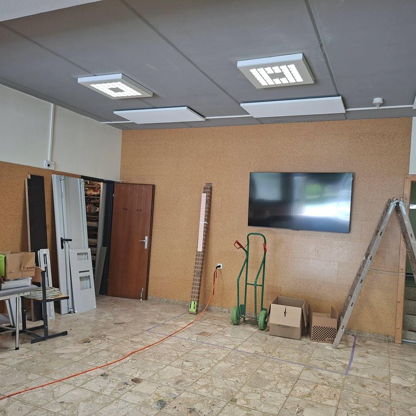 Ein leerer Raum mit braunen Wänden hat einen Flachbildfernseher an der Wand montiert, Lagerboxen, einen Trolley und eine Leiter. Eine Tür und Regale befinden sich auf der linken Seite, mit einem Tisch auf der linken Seite.