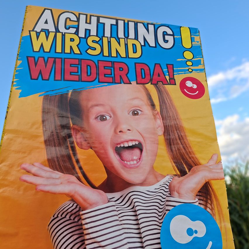 Ein farbenfrohes Poster mit einem jungen Mädchen, das seinen Mund öffnet und die Hände hebt, trägt ein gestreiftes Shirt, vor einem gelben Hintergrund mit blauen Akzenten und dem Text 'Achtung Wir sind wieder da!'.