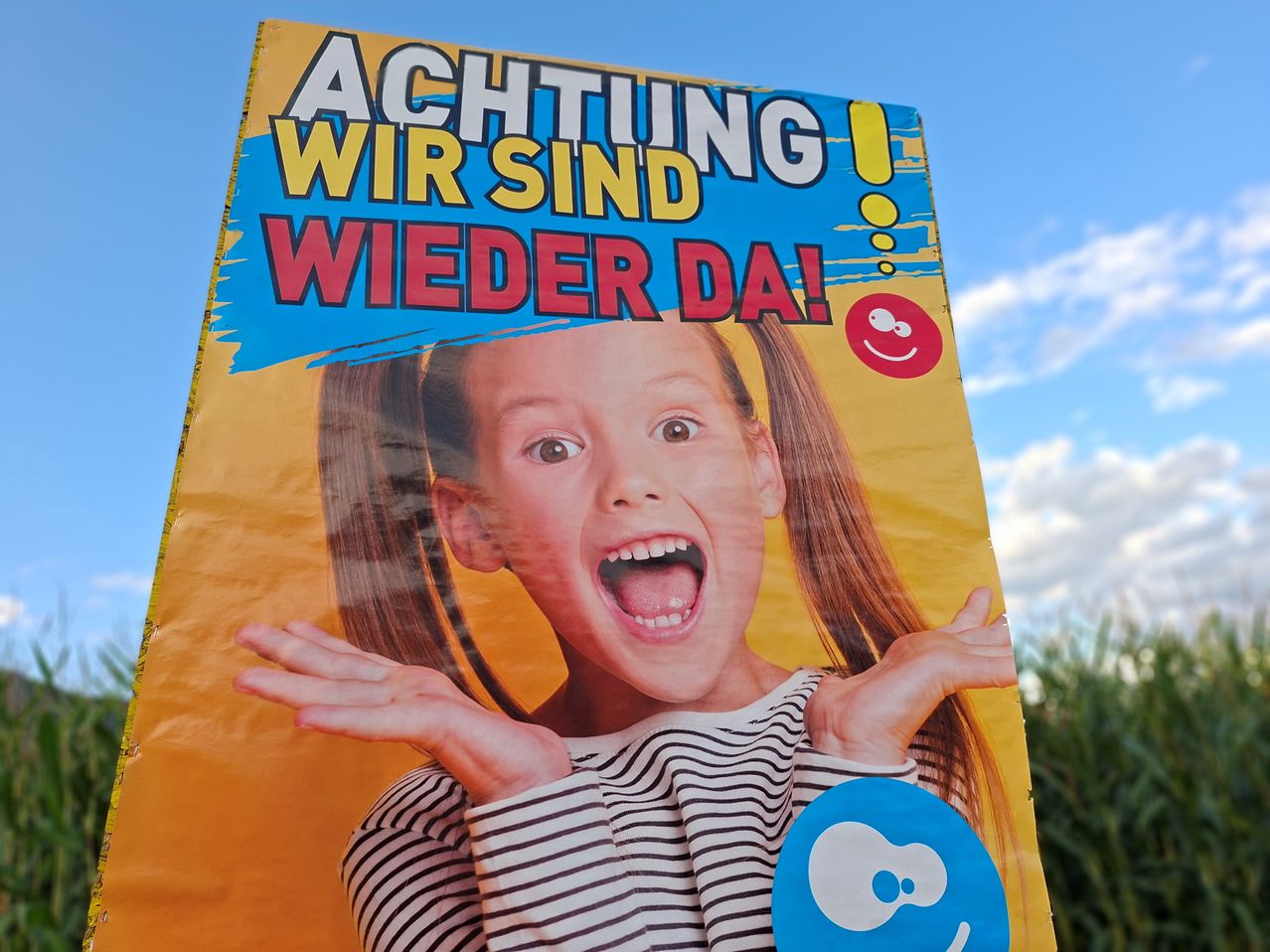 Ein farbenfrohes Poster mit einem jungen Mädchen, das seinen Mund öffnet und die Hände hebt, trägt ein gestreiftes Shirt, vor einem gelben Hintergrund mit blauen Akzenten und dem Text 'Achtung Wir sind wieder da!'.