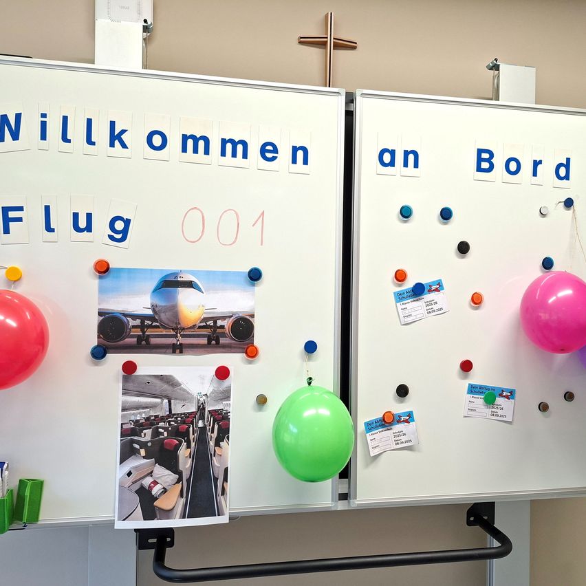 Ein Whiteboard mit blauen Buchstaben, die 'Willkommen Flug 001' schreiben, und Bildern eines Flugzeugs. Bunte Luftballons und Namensschilder sind befestigt.