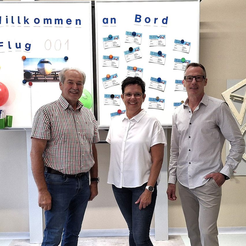 Drei Personen stehen neben einer weißen Tafel mit 'Willkommen an Bord' und 'Flug 001'. An der Tafel sind mehrere Karten befestigt. Die Personen lächeln.