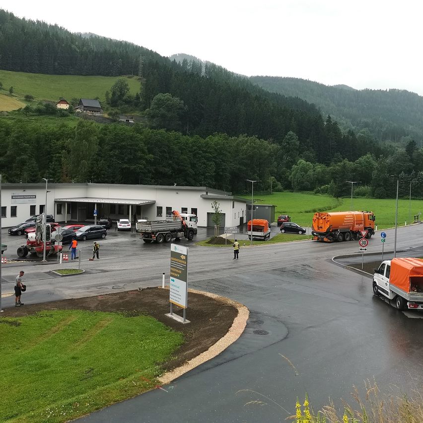 Ein belebter Bereich mit einem großen weißen Gebäude, mehreren Autos, Lastwagen und Maschinenfahrzeugen. Menschen arbeiten in dem Bereich. Grüne Hügel und Bäume sind im Hintergrund.