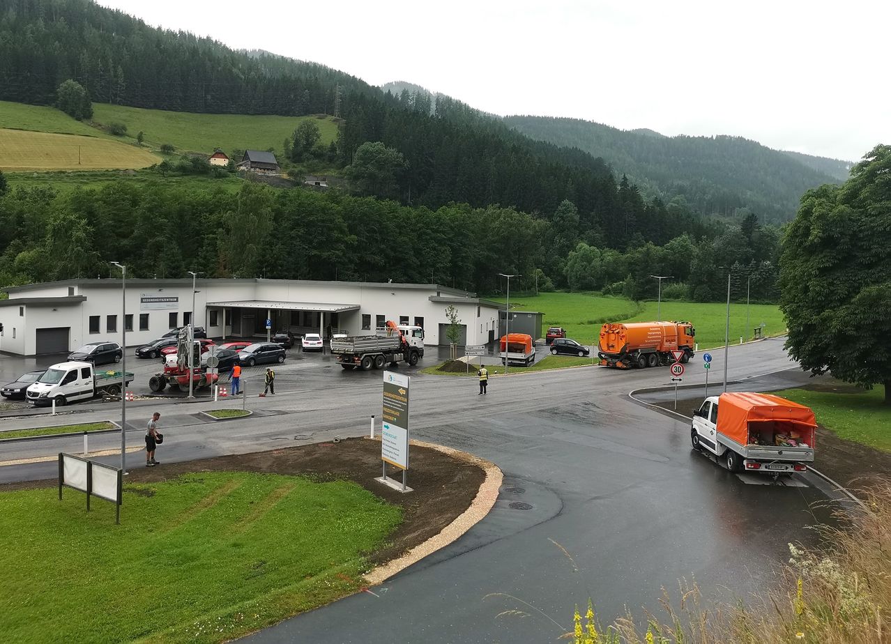 Ein belebter Bereich mit einem großen weißen Gebäude, mehreren Autos, Lastwagen und Maschinenfahrzeugen. Menschen arbeiten in dem Bereich. Grüne Hügel und Bäume sind im Hintergrund.