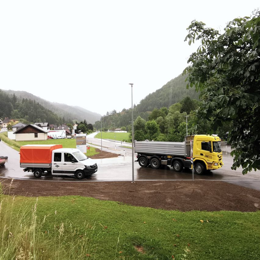 Zwei Lastwagen fahren auf einer Straße mit Häusern und Bergen im Hintergrund. Ein Lastwagen ist gelb mit einem silbernen Container, und der andere ist weiß mit einer orangefarbenen Abdeckung.