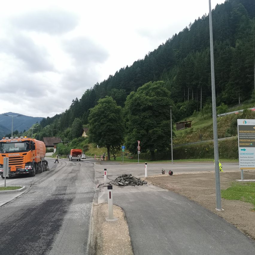 Eine Baustelle mit einem orangefarbenen Lkw und einer Person, die geht. Es gibt Haufen von Kies auf der Seite. Die Szene umfasst einen Berg, ein Haus und Straßenlaternen.