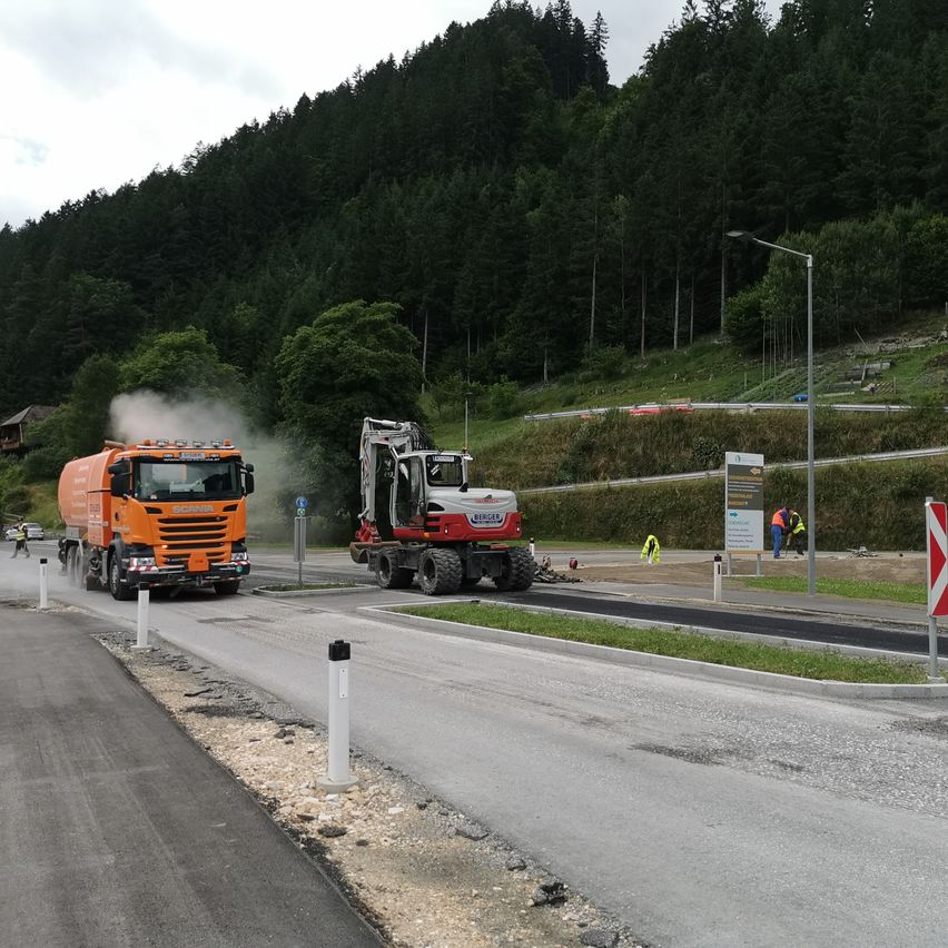 Eine Baustelle mit einem orangefarbenen Lastwagen und einem rot-weißen Bagger auf der Straße. Arbeiter in Sicherheitswesten sind anwesend, und eine Straßenlaterne steht an der Seite.