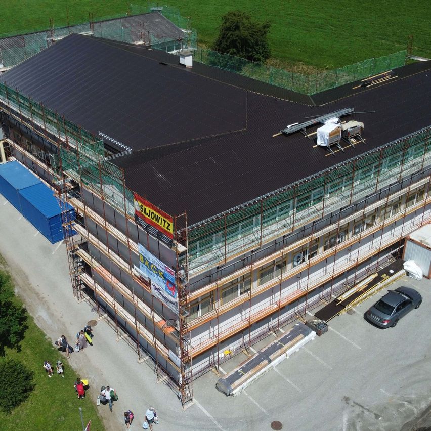 Ein Luftbild eines Gebäudes im Bau. Das Dach ist mit schwarzem Dachmaterial bedeckt, und Gerüst umgibt die Struktur. Ein Auto ist in der Nähe geparkt.