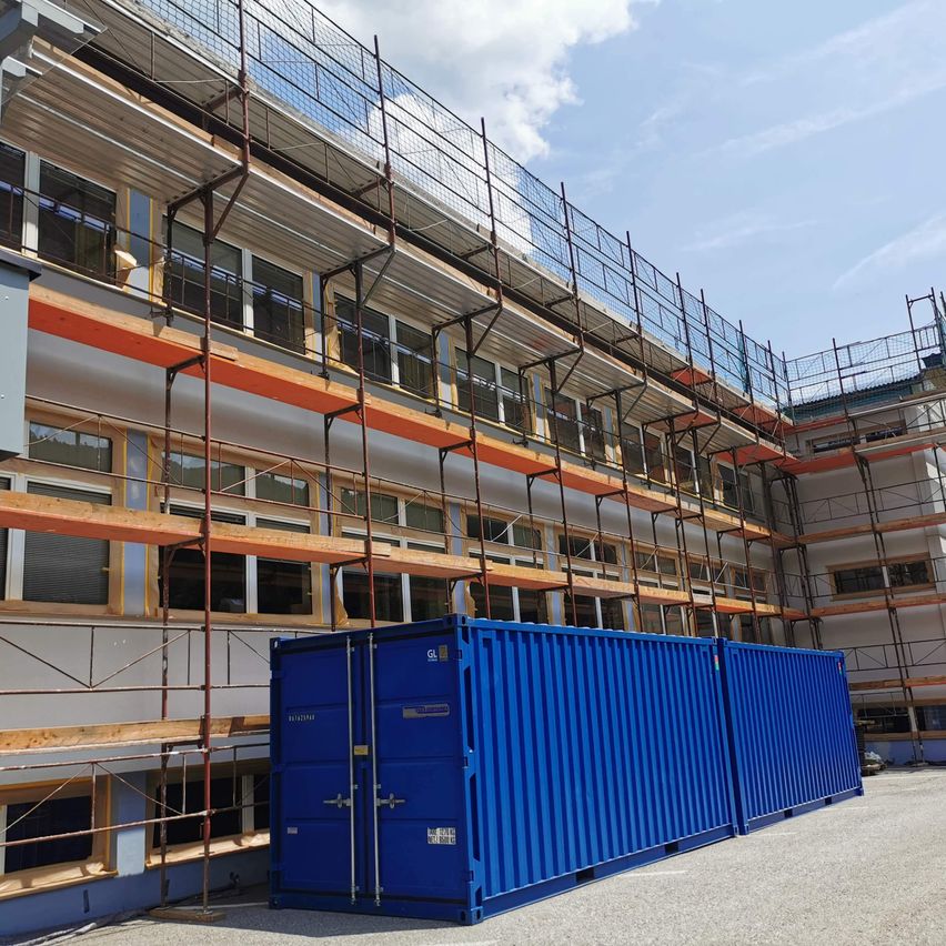 Ein Gebäude im Bau mit Gerüsten und zwei blauen Containern davor. Der Himmel ist blau mit einigen Wolken.