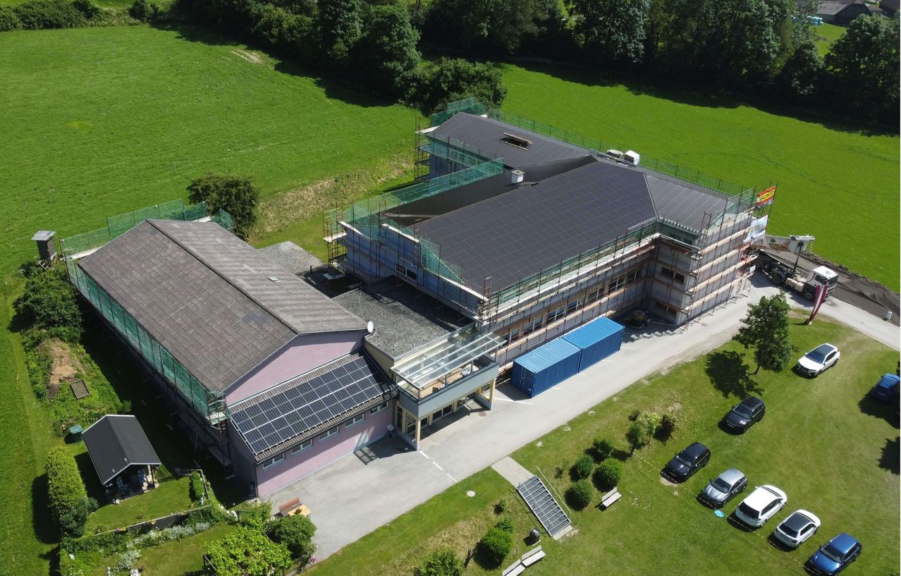 Ein Luftbild eines Gebäudes mit Solarpanelen auf dem Dach im Bau. In der Nähe sind Autos geparkt und das Gebiet ist von Grünflächen umgeben.