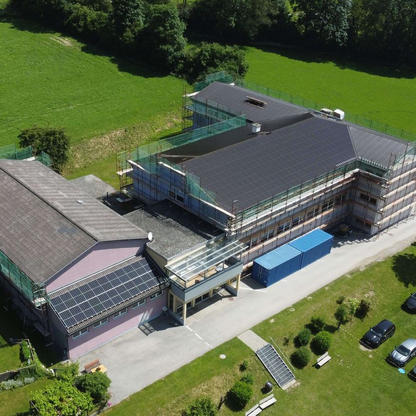 Ein Luftbild eines Gebäudes mit Solarpanelen auf dem Dach im Bau. In der Nähe sind Autos geparkt und das Gebiet ist von Grünflächen umgeben.