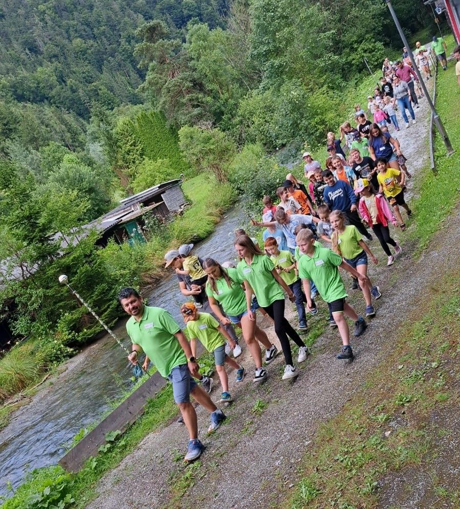 Eine Gruppe von Menschen in grünen Shirts läuft auf einem Pfad neben einem Fluss mit Bäumen und einem kleinen Haus im Hintergrund.