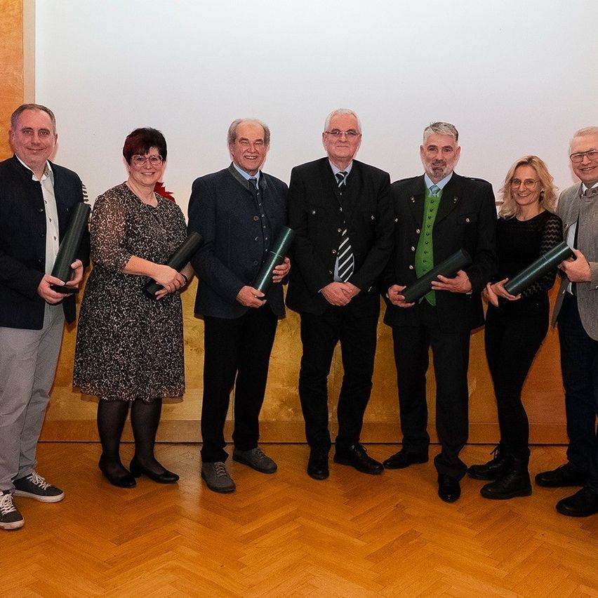 Eine Gruppe von Personen steht vor einem weißen Hintergrund. Sie halten aufgerollte Schriftrollen. Die Leute sind in formeller Kleidung gekleidet. Sie lächeln.