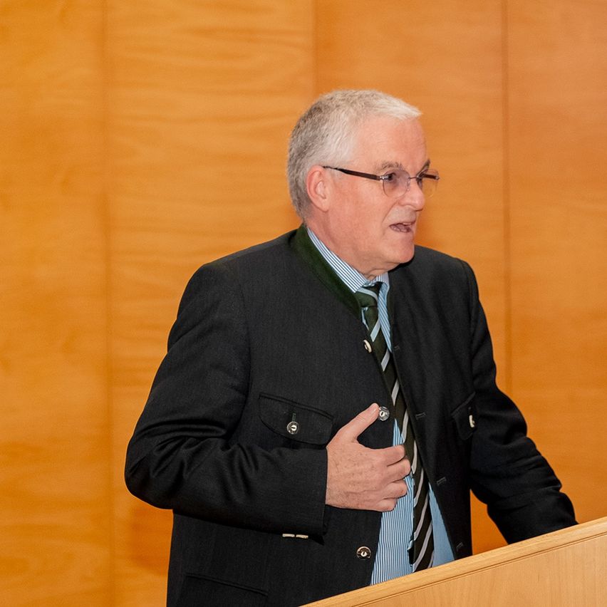 Ein älterer Mann steht an einem Podium, trägt einen schwarzen Mantel, ein blaues Hemd und eine gestreifte Krawatte. Er scheint eine Rede zu halten.