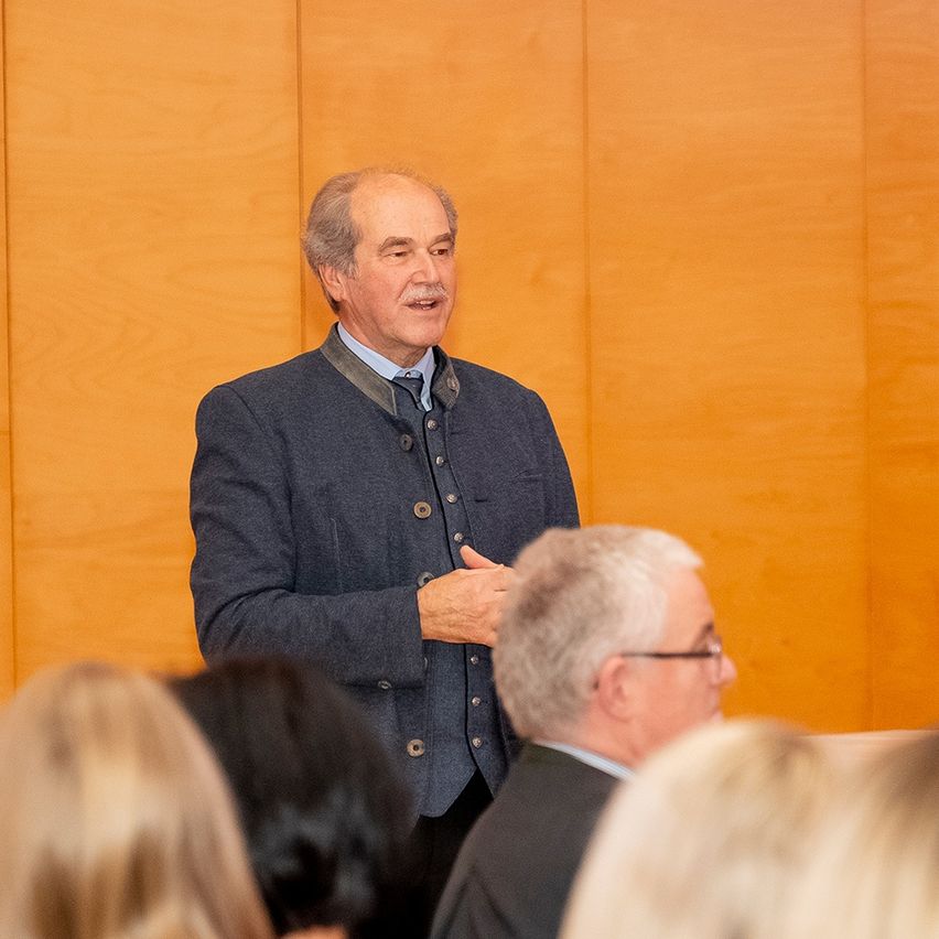 Ein Mann in einem grauen Anzug spricht zu einer Gruppe von Menschen in einem Konferenzraum.