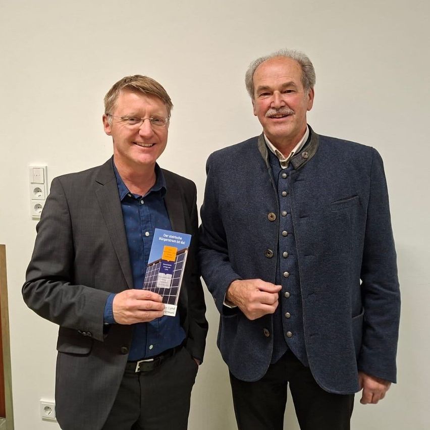 Zwei Männer stehen nah beieinander in einer Innenraumsituation. Der Mann auf der linken Seite hält ein Buch mit dem Titel 'Unsere elektrischen' und trägt eine Brille und einen Gürtel. Der Mann auf der rechten Seite lächelt und trägt einen marineblauen Blazer mit Knöpfen. Hinter ihnen ist eine weiße Wand mit Steckdosen.