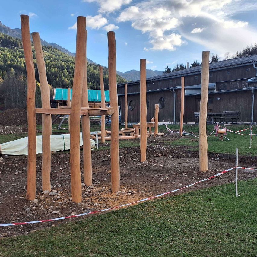Ein Baugelände mit Holzpfosten ist auf einem Rasenbereich errichtet, mit einem Gebäude und Bergen im Hintergrund.