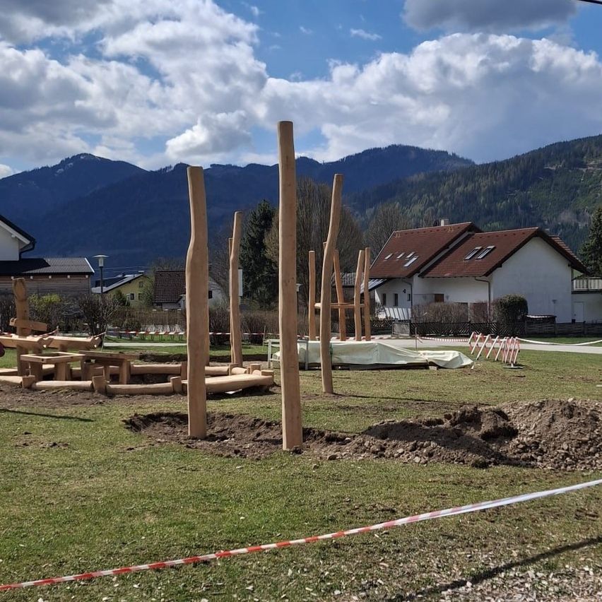 Ein Spielplatz im Bau in einem Rasenfeld mit Holzpfosten, einem weißen Haus mit braunem Dach, Bergen im Hintergrund und einem bewölkten Himmel.