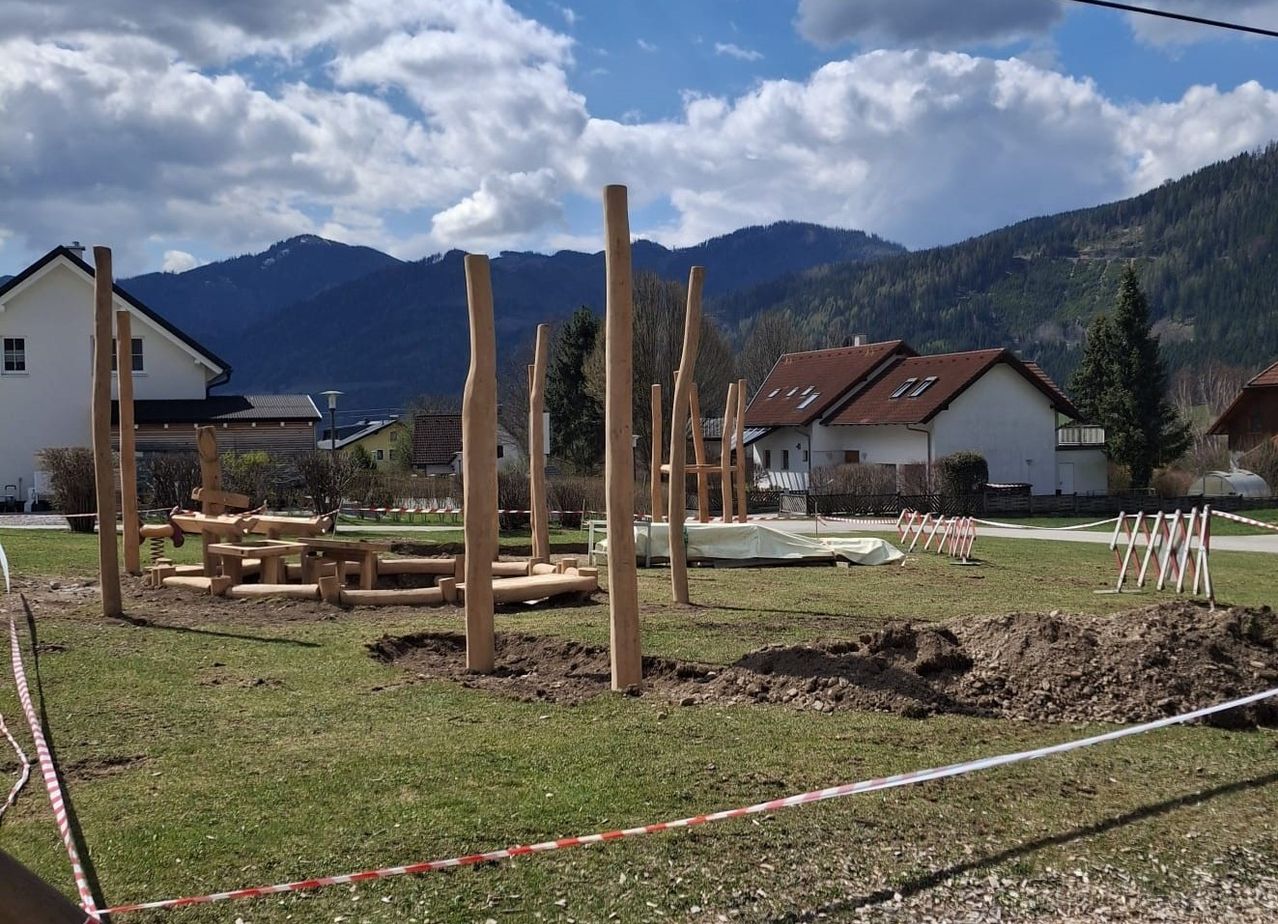 Ein Spielplatz im Bau in einem Rasenfeld mit Holzpfosten, einem weißen Haus mit braunem Dach, Bergen im Hintergrund und einem bewölkten Himmel.