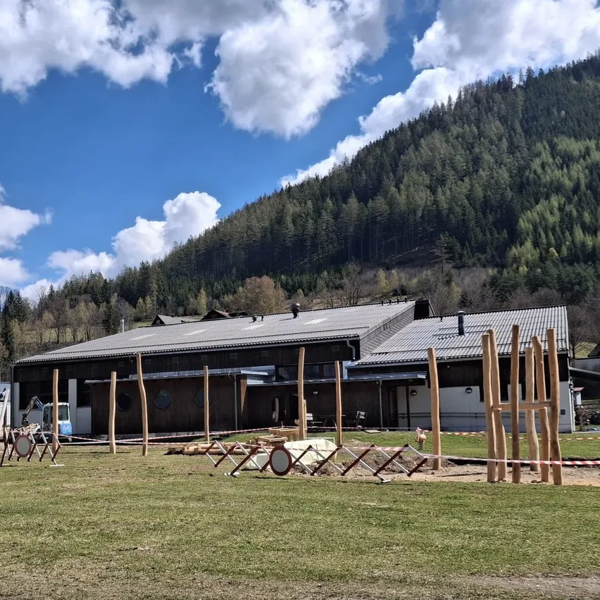 Ein großes Gebäude mit einem flachen Dach und großen Fenstern steht vor einem Berg. Holzpfosten sind im Gras aufgestellt.