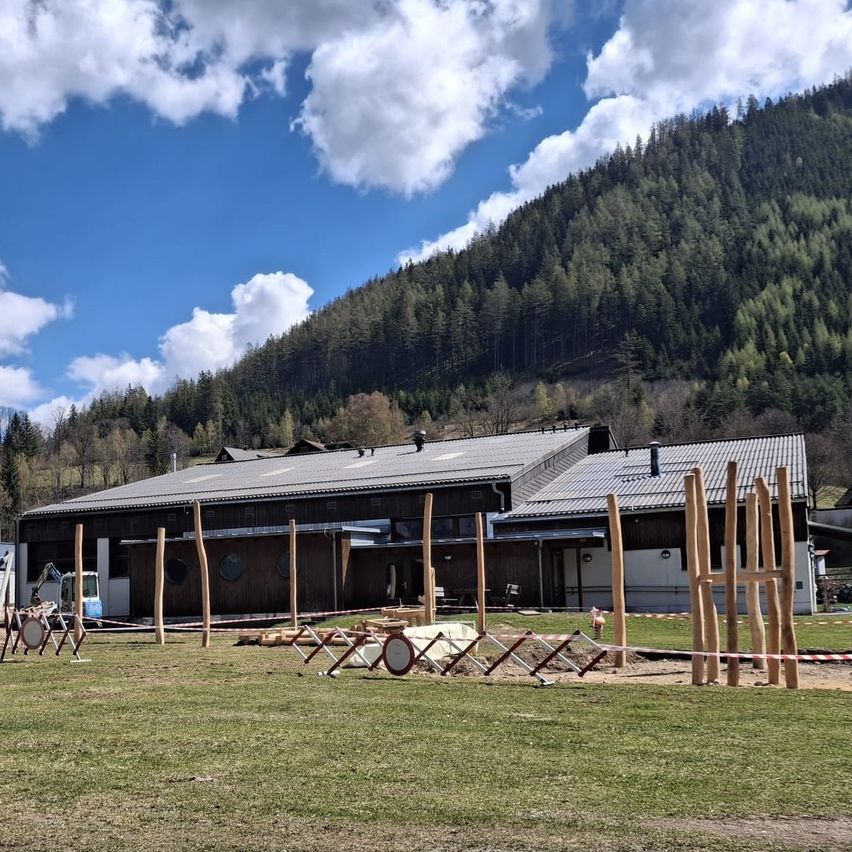 Ein großes Gebäude mit einem flachen Dach und großen Fenstern steht vor einem Berg. Holzpfosten sind im Gras aufgestellt.