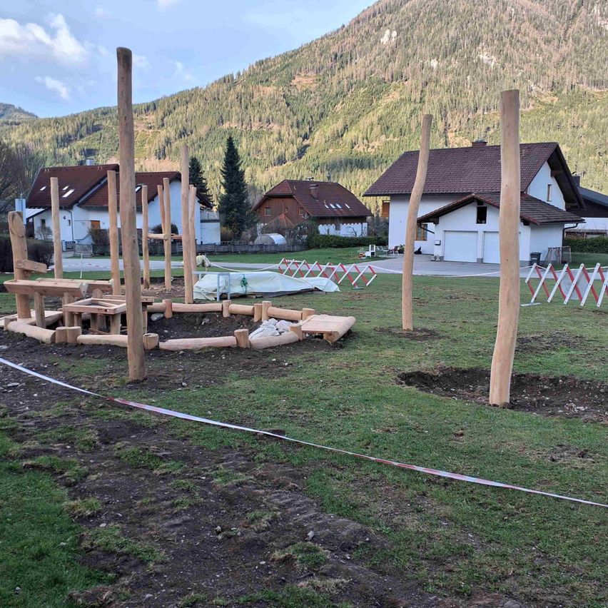 Ein Rasenbereich mit einer Holzstruktur, umgeben von Holzpfosten. Dahinter befinden sich drei Häuser mit braunen Dächern. In der Ferne erhebt sich ein Berg, der von Bäumen bedeckt ist.