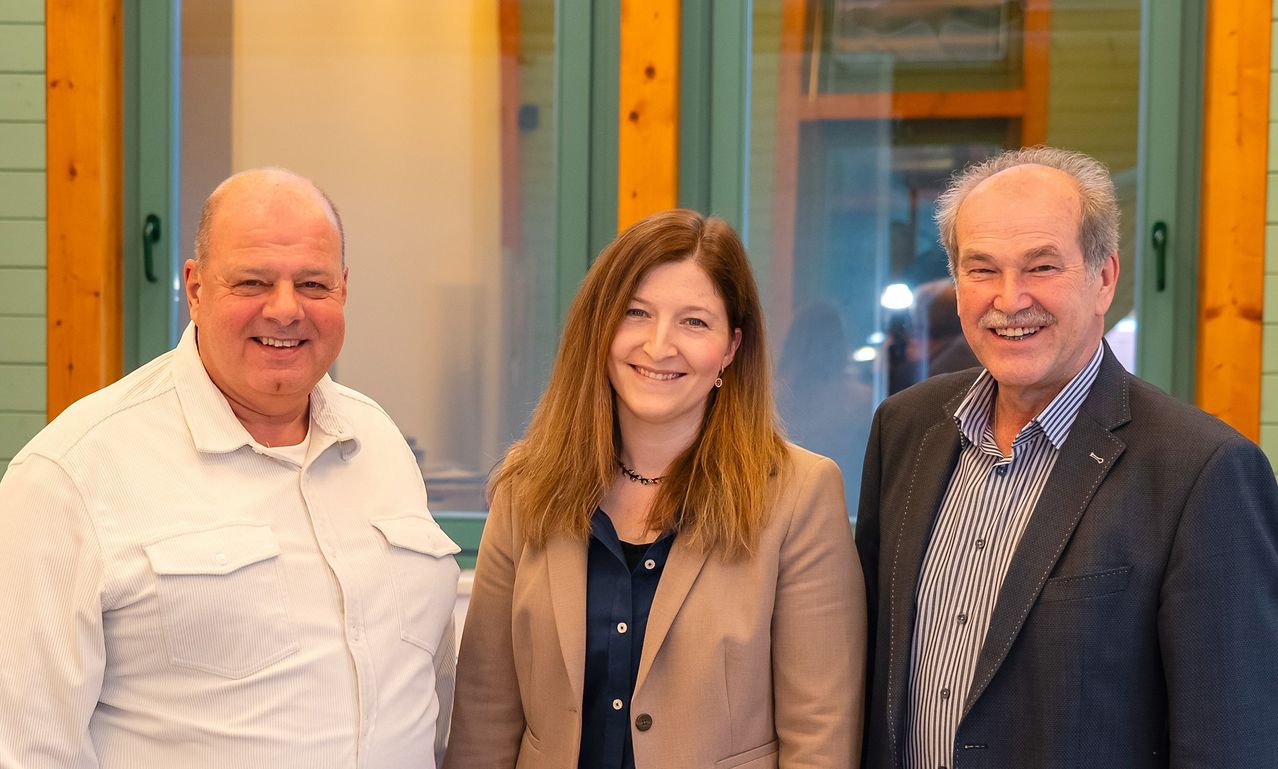 Drei Menschen stehen eng beieinander und lächeln für ein Foto. Ein Mann in Weiß steht links, eine Frau in einer braunen Jacke in der Mitte und ein Mann in einem gestreiften Hemd rechts. Dahinter befindet sich eine Glaswand, die einen Raum mit Holzrahmen reflektiert.