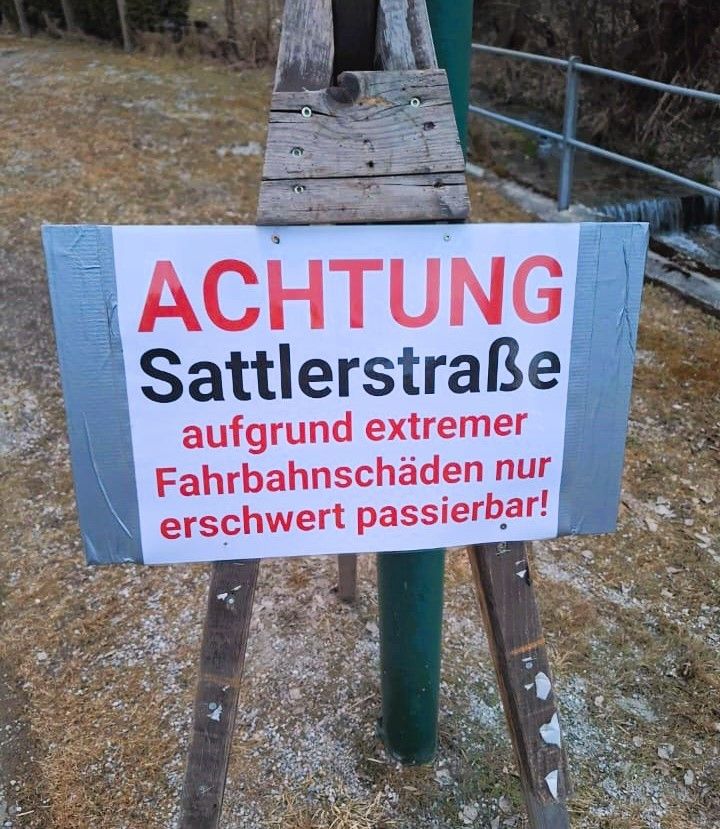 Ein Schild auf einem Holzständer steht mit roten Buchstaben 'ACHTUNG Sattlerstrasse', was auf extreme Bahnschäden hinweist, und ist nur schwer passierbar.