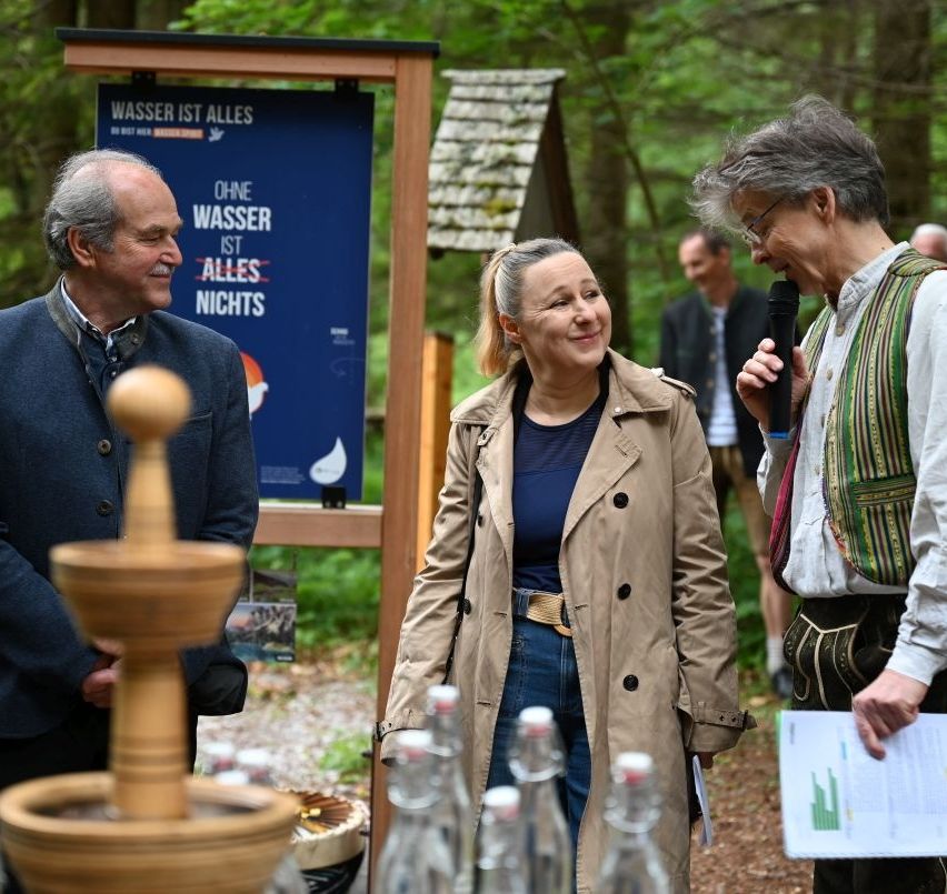 Eine Gruppe von Menschen steht in einer Waldumgebung, eine Frau in einem Trenchcoat lächelt und schaut einen Mann an, der ein Mikrofon hält. Hinter ihnen steht ein Schild mit der Aufschrift 'Wasser ist alles' und eine Holzstruktur ist sichtbar.