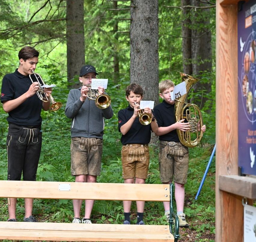 Vier Jungen in traditioneller deutscher Kleidung stehen in einem Wald, jeder hält ein Blechblasinstrument und liest von einem Notenblatt.