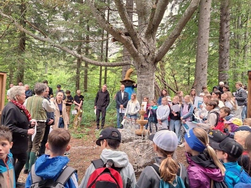 Eine Gruppe von Menschen versammelt sich in einem Wald, einige stehen und andere sitzen auf dem Boden. Ein Mann steht vor einem Baum mit einem großen Hut darauf. Ein Brunnen steht vor dem Baum. Einige Leute halten Papiere und Bücher.