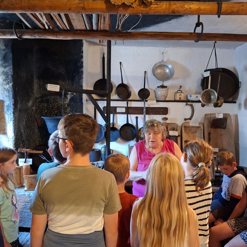 Eine ältere Frau steht vor einer Gruppe von Kindern und erklärt etwas in einer rustikalen Küche, in der verschiedene Töpfe und Pfannen an der Wand hängen.