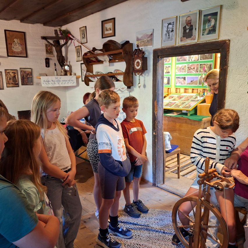 Eine Gruppe von Kindern und Erwachsenen versammelt sich in einem Raum mit Holzböden und -wänden, beobachtet eine Spinnmaschine und eine Frau.