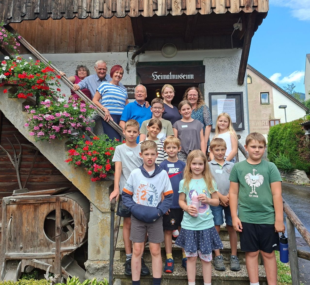 Eine Gruppe von Menschen, einschließlich Erwachsener und Kindern, steht auf den Stufen eines Gebäudes mit einem Schild, das 'Seimatmuseum' besagt. Sie lächeln und posieren für ein Foto.