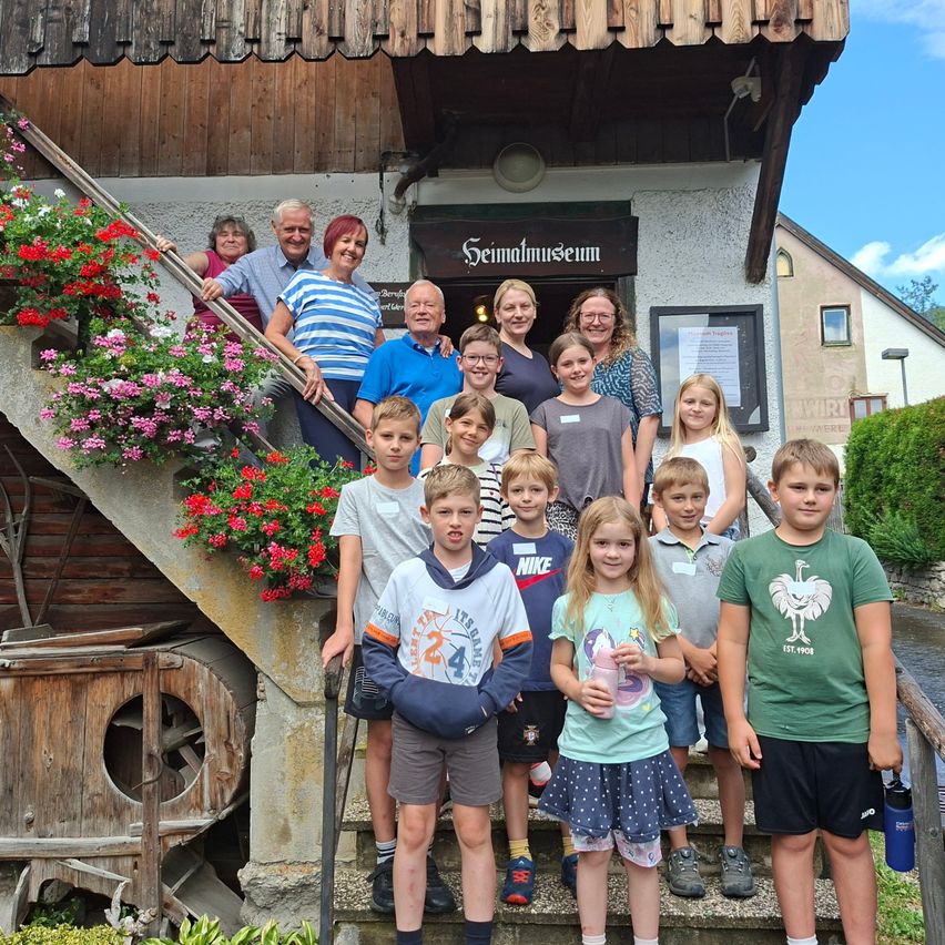 Eine Gruppe von Menschen, einschließlich Erwachsener und Kindern, steht auf den Stufen eines Gebäudes mit einem Schild, das 'Seimatmuseum' besagt. Sie lächeln und posieren für ein Foto.