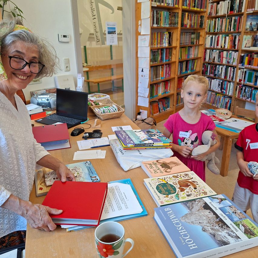 Eine ältere Frau mit Brille und Kopfband lächelt, während sie ein rotes Buch in einer Bibliothek hält. Ein junges Mädchen in einem rosa Shirt steht neben ihr und hält ein Kuscheltier. Ein Schreibtisch ist mit Büchern, einem Laptop und einer Tasse gefüllt.