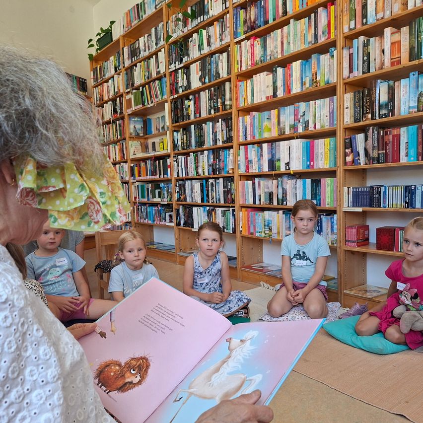 Eine ältere Frau liest ein Bilderbuch für eine Gruppe junger Kinder, die in einem Kreis in einer Bibliothek sitzen.