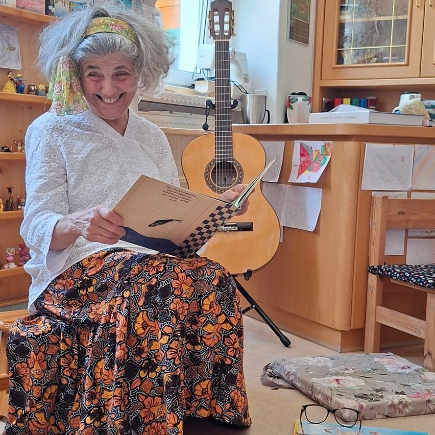 Eine Frau sitzt auf einem Hocker und liest ein Buch, hält eine Gitarre hinter sich, in einem Raum mit Regalen und einem Schreibtisch.