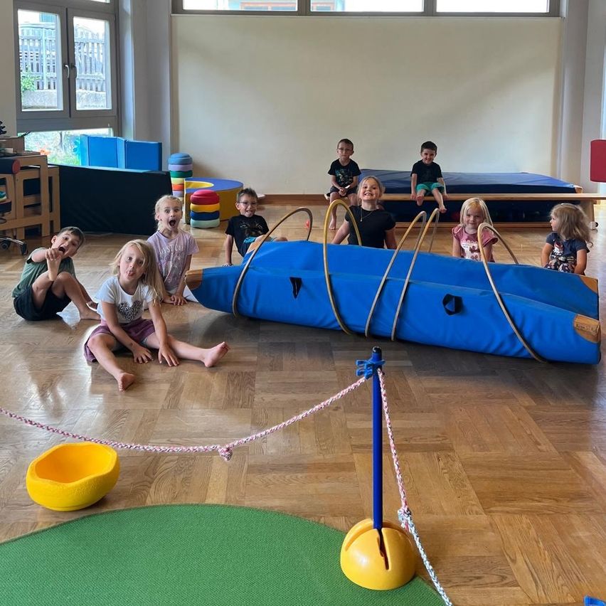 Eine Gruppe von Kindern spielt in einem Raum mit einem blauen aufblasbaren Tunnel und verschiedenen Spielzeugen. Einige sitzen auf dem Boden, während andere stehen.