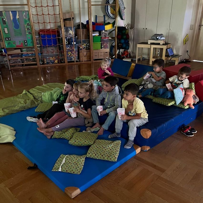 Eine Gruppe von Kindern sitzt auf einer blauen Matte in einem Raum, hält Tassen und Snacks und einige tragen Brillen.
