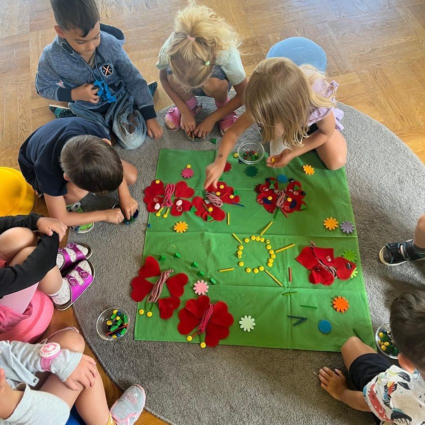 Eine Gruppe von Kindern sitzt auf dem Boden um eine grüne Matte herum und ist in einer Bastelaktivität verwickelt. Sie verwenden verschiedene Materialien wie Filzblumen, Schnüre und Perlen, um etwas zu kreieren.
