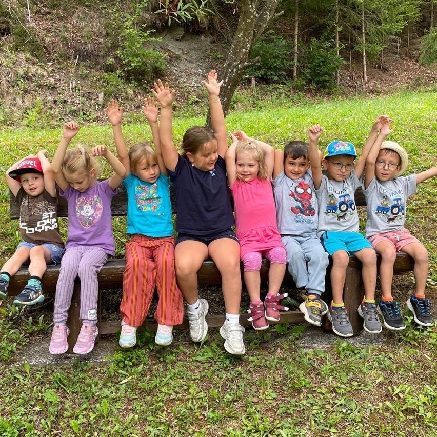 Eine Gruppe von Kindern sitzt auf einer Holzbank in einem Park, alle mit erhobenen Händen. Sie tragen Freizeitkleidung und Turnschuhe. Hinter ihnen befindet sich ein Grasbereich mit Bäumen und einem Pfad.