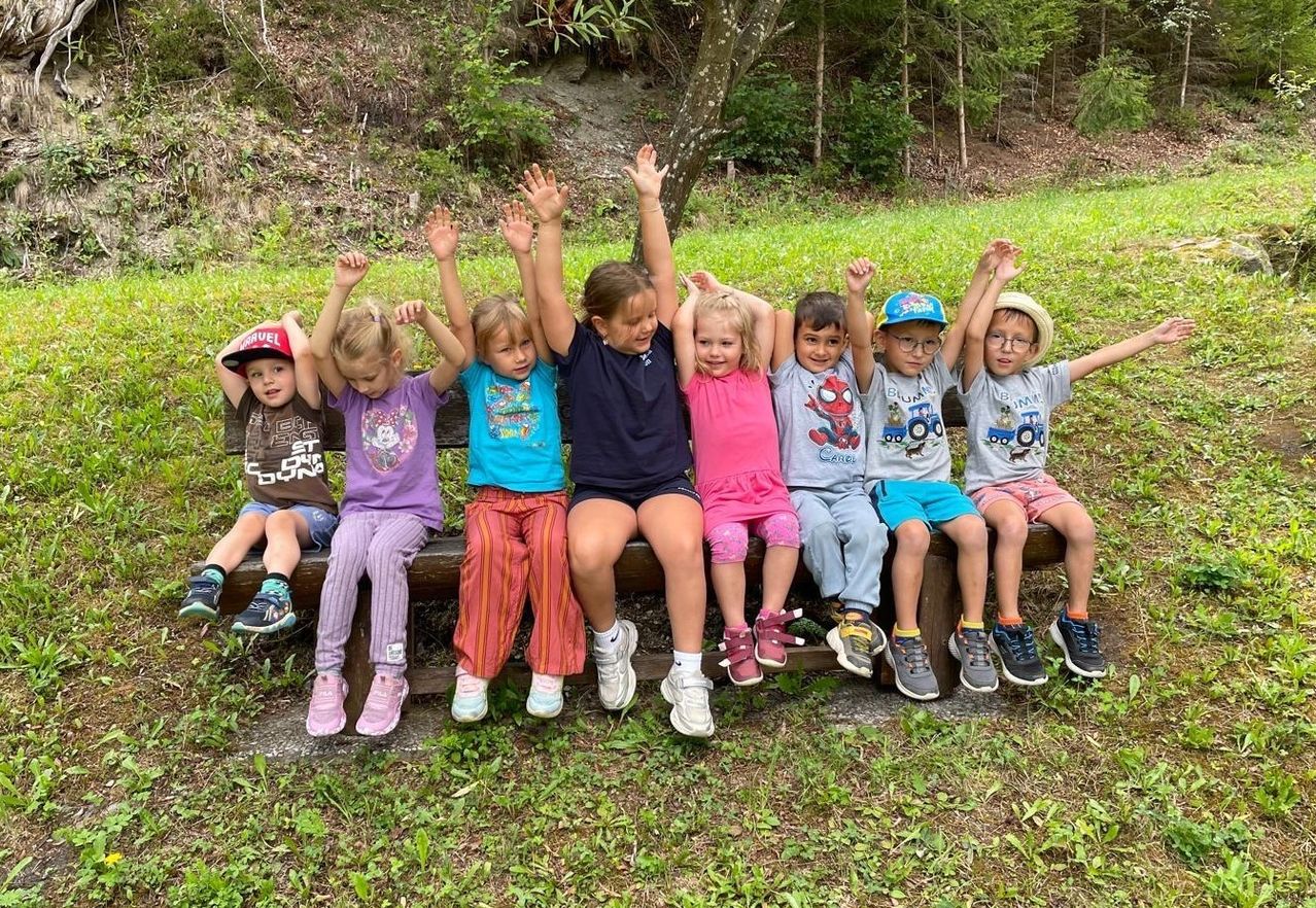 Eine Gruppe von Kindern sitzt auf einer Holzbank in einem Park, alle mit erhobenen Händen. Sie tragen Freizeitkleidung und Turnschuhe. Hinter ihnen befindet sich ein Grasbereich mit Bäumen und einem Pfad.