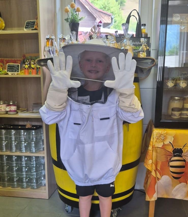 A young boy in a beekeeper's suit stands in front of a yellow barrel with bee decorations, smiling and posing for a photo.