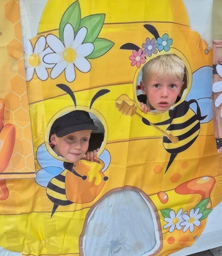 Zwei kleine Jungen schauen aus einem bienenthematischen Spielzelt, einer hält ein Honigglas und der andere ein Bienenwaben.