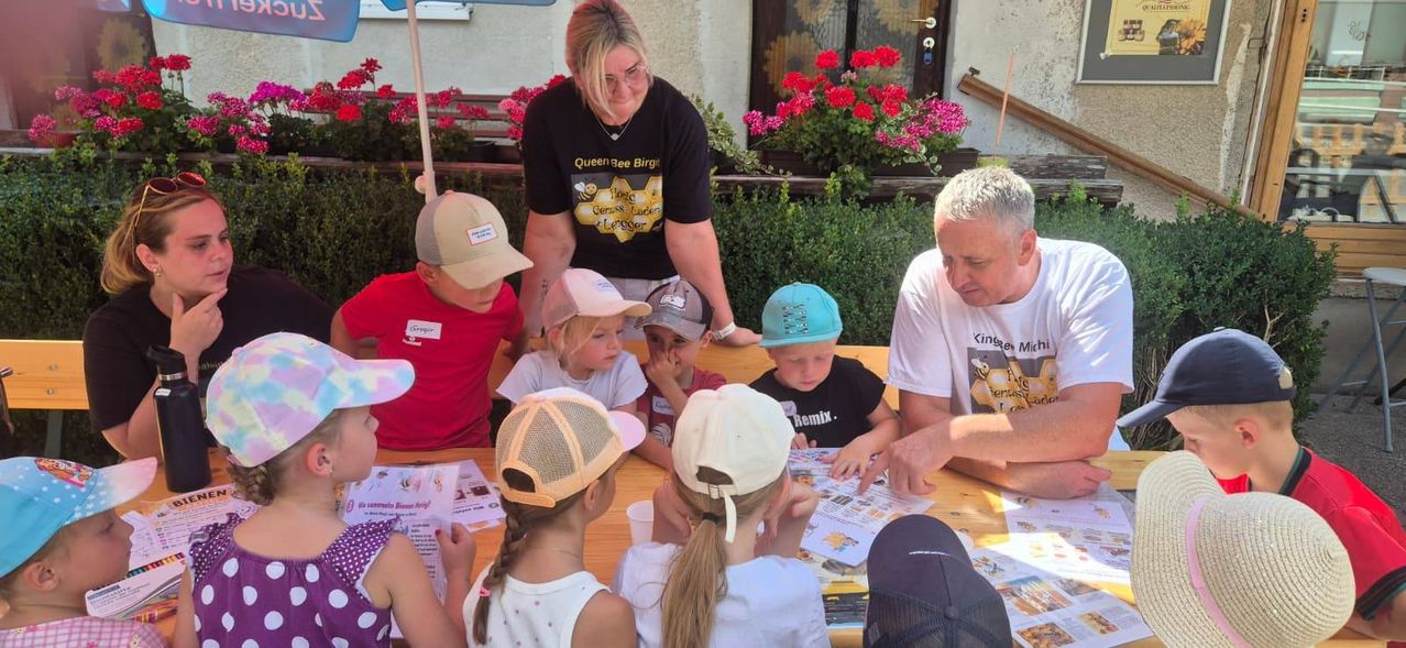 A group of children and two adults are sitting around a table with papers and cups, with one adult wearing a bee-themed shirt.