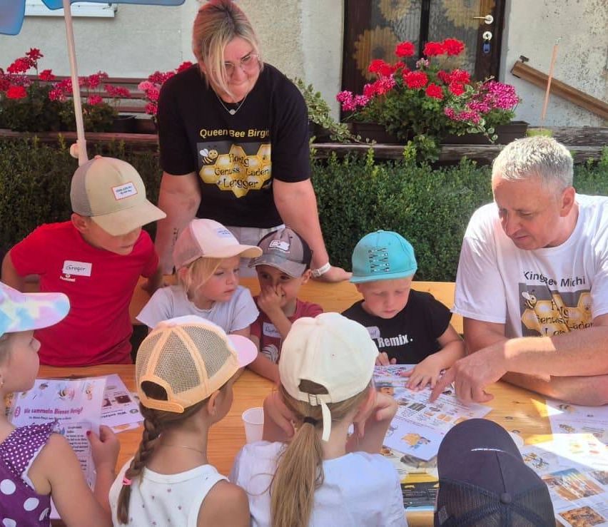Eine Gruppe von Kindern und zwei Erwachsenen sitzen um einen Tisch mit Papieren und Bechern, wobei einer der Erwachsenen ein bienenthematisches Shirt trägt.